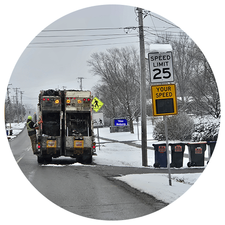 Garbage truck on snowy road, passing speed limit sign showing 25 mph. Worker in high-visibility clothing stands by the truck. Snow-laden trees and bins line the street.