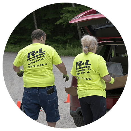 Two workers in yellow "R&L Rubbish" shirts load cardboard into a red SUV on a forest-lined road, with traffic cones marking the area.